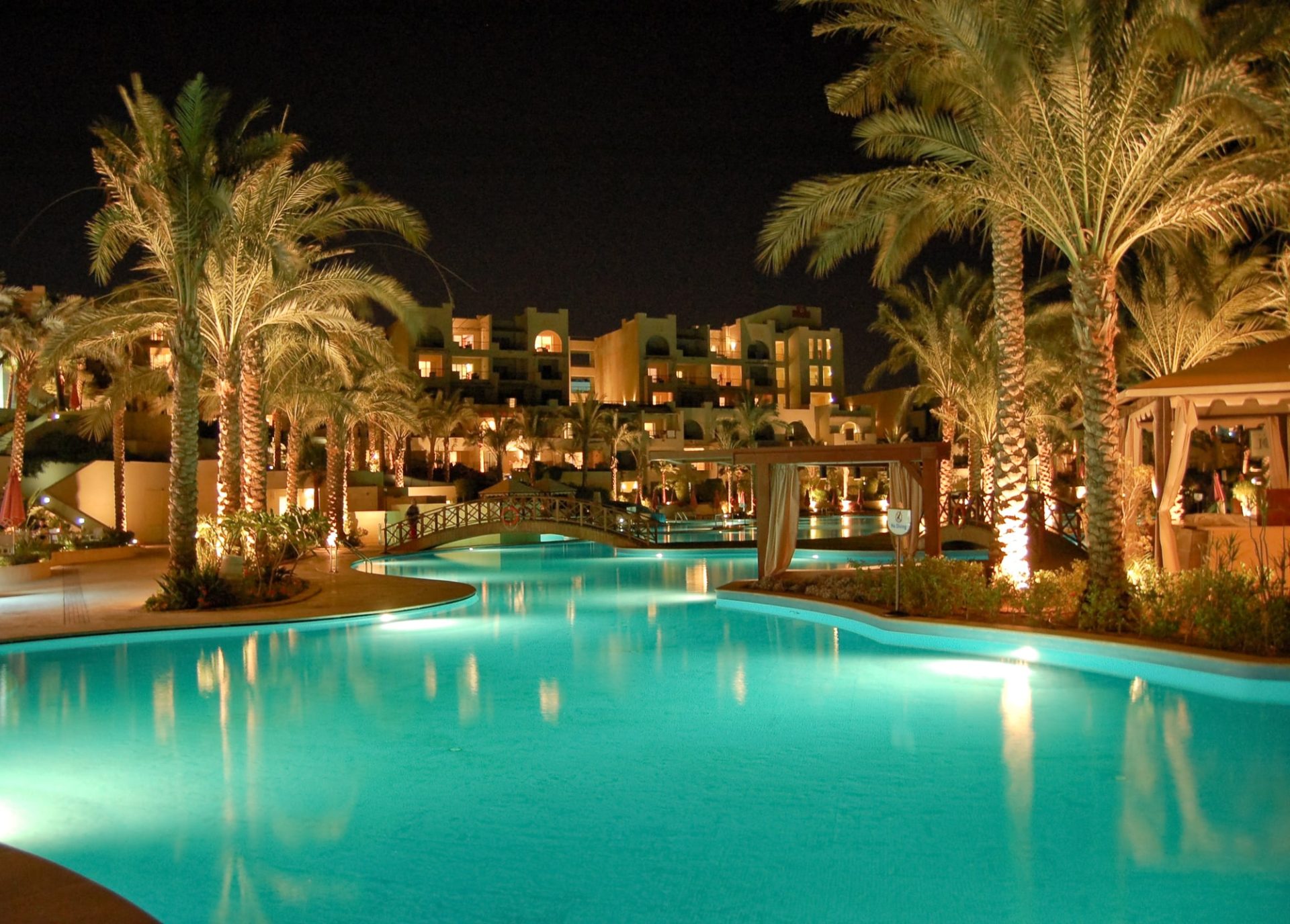 A luxurious hotel swimming pool at night with palm trees surrounding it.