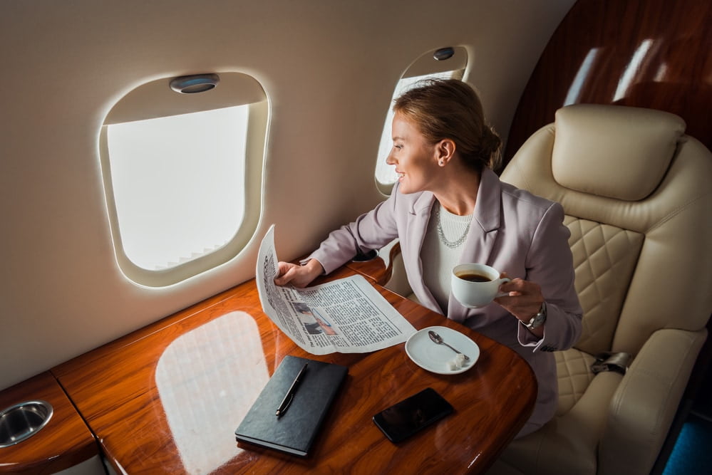 Cheerful businesswoman holding cup and business newspaper near smartphone with blank screen in private plane — Photo