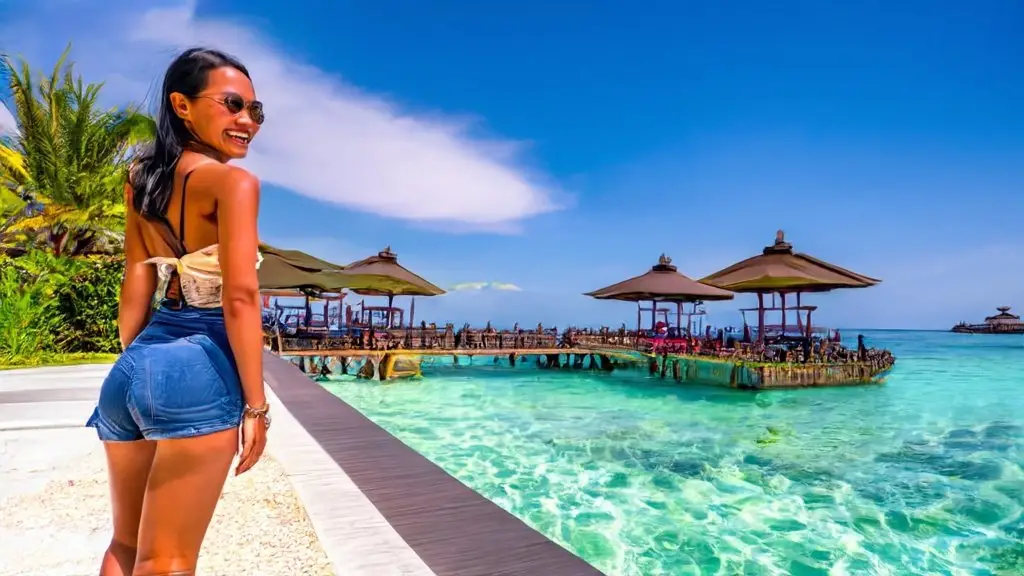A woman smiles by a poolside walkway with tropical thatched-roof structures over clear turquoise waters in the background during her cheap luxury vacation.