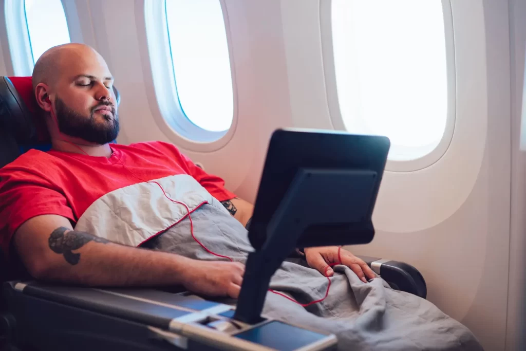 A man with a closely shaved head, wearing a red t-shirt and sporting a tattoo on his arm, is sleeping in a first-class airplane seat with a blanket on his lap, next to a window.