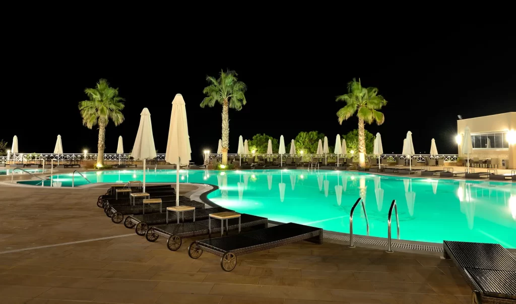 A night-time view of a serene Daios Cove Luxury Resort swimming pool illuminated by ambient lights, surrounded by palm trees and loungers under closed white umbrellas.