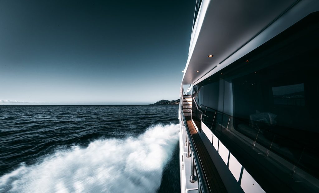 Side view of a Ritz-Carlton yacht moving at high speed on the ocean, with water splashing and a clear blue sky in the background, illustrating a sense of luxury with Ritz-Carlton Yachting.