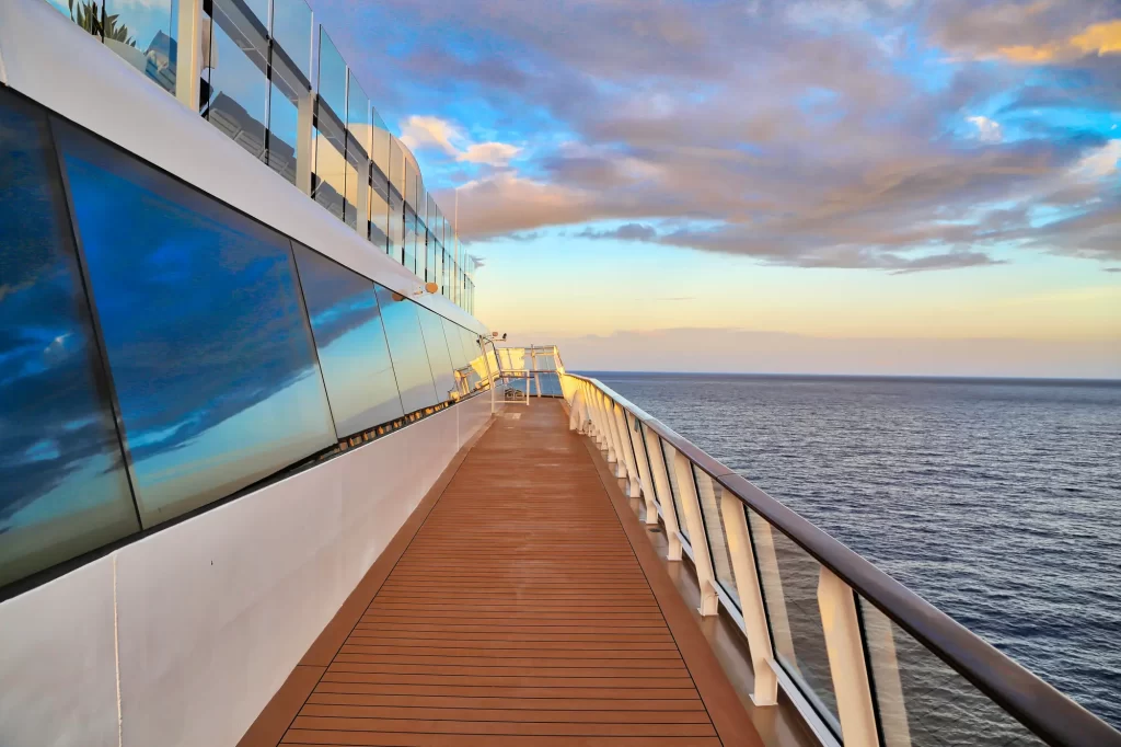 A view from the deck of a Ritz-Carlton Yachting cruise ship at sunset, showcasing the reflective blue glass balcony, wooden floor, and the vast ocean extending into the horizon under a