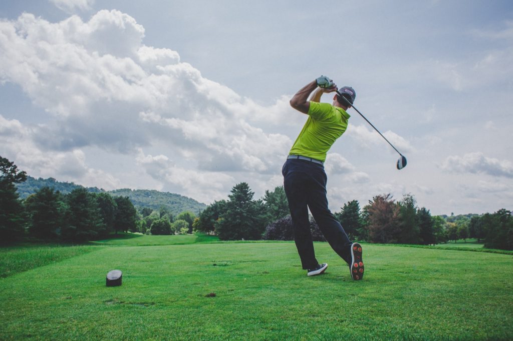 A golfer in a yellow shirt and black pants swings a club on a tee box at one of the best golf resorts in Central America, surrounded by lush greenery under a partly cloudy sky. A